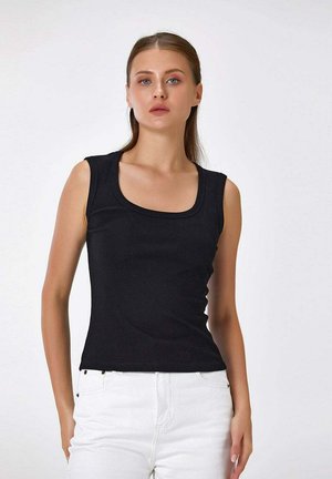 Jeune femme portant un haut noir sans manches et un pantalon blanc, debout devant un fond clair uni, faisant face à l’avant.
