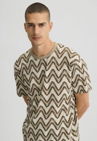 Camiseta de manga corta en beige con un patrón de zigzag marrón, con cuello redondo y textura de algodón suave.
