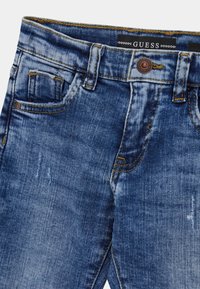 Modré vybledlé džíny z denimu s předními kapsami, zapínáním na knoflík, poutky na pásek a viditelnou značkovou štítkem Guess uvnitř pásku.