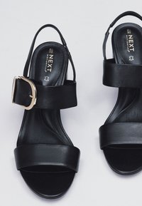 Schwarze Sandaletten mit Absatz, weitem Vorderzriemen und dünnem Knöchelriemen. Mit einem goldfarbenen Schnallenakzent. Glatte Lederstruktur.