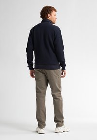 Granatowy sweter o fakturze z wysokim kołnierzem, zestawiony z jasnokawowymi spodniami. Model stoi tyłem, prezentując swobodny krój i design.