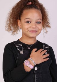 Zilveren horloge met roze wijzerplaat en band, zilveren hartvormige oorbellen, en een bijpassende ketting en armband op een meisje in een zwarte shirt en overall.