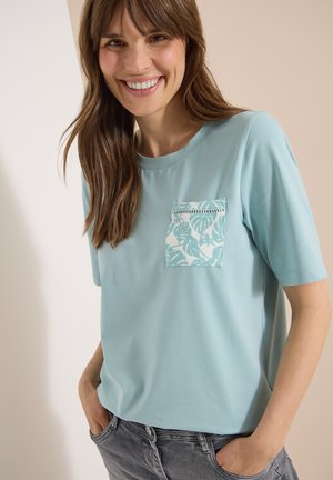 Kvinde med langt brunt hår smiler iført en lyseblå t-shirt med korte ærmer, med et mønstret brystlomme og grå jeans.