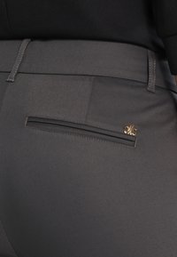 Mörkgrå byxor med en slät yta, med en bakficka med vertikal öppning, accentuerad av en liten guldfärgad logotypdetalj.