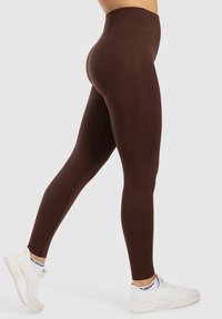 Leggings marroni a vita alta realizzati in un tessuto elasticizzato, progettati con una texture liscia e dettagli di cucitura. Abbinati a scarpe da ginnastica bianche.