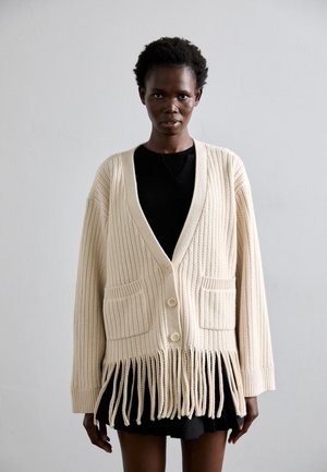 Krémový rebrovaný cardigan s frayed lemom, má výstrih do V, dve predné vrecká a zapínanie na gombíky, vrstvený cez čierny top.