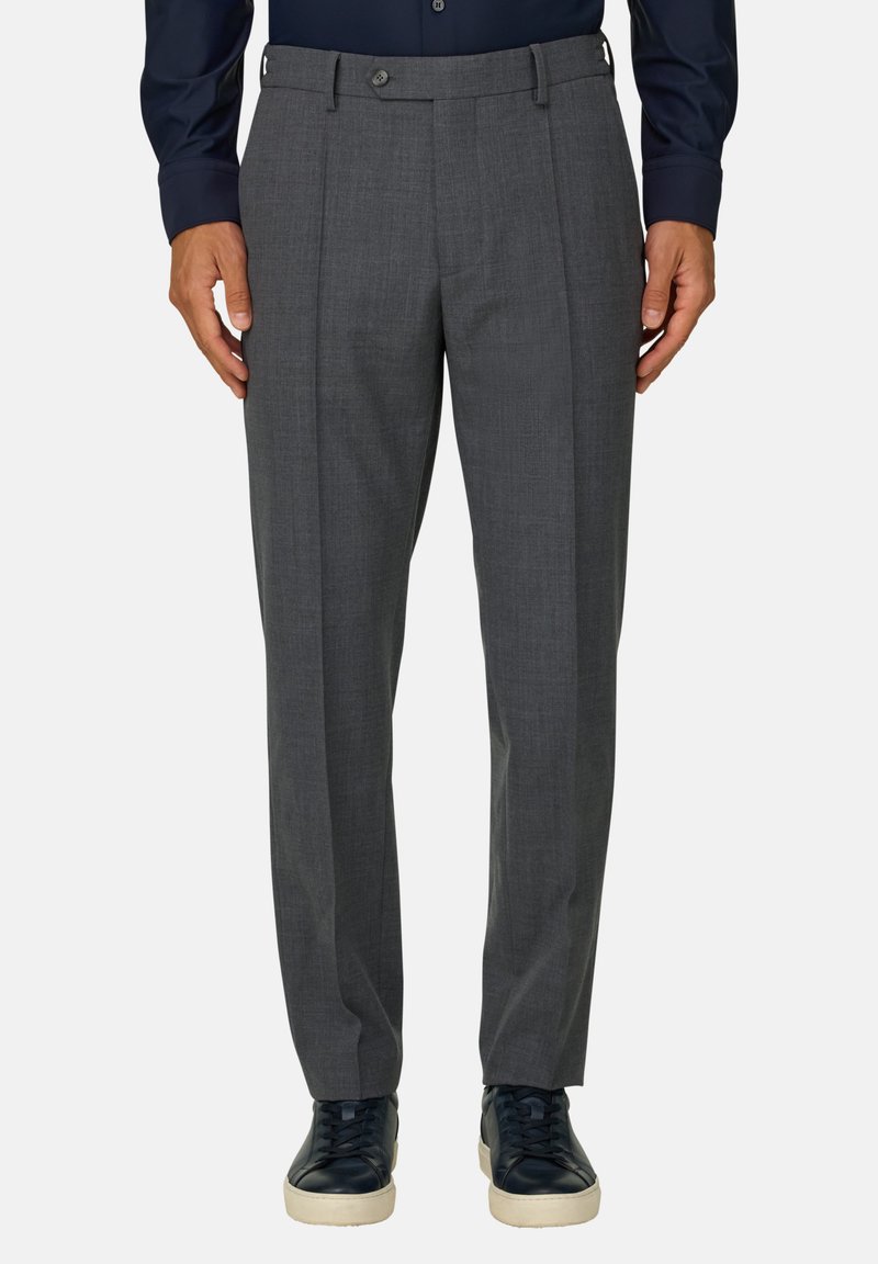 Homme portant un pantalon gris sur mesure, une chemise bleu marine foncé et des baskets noires avec des semelles blanches, debout devant un fond uni.