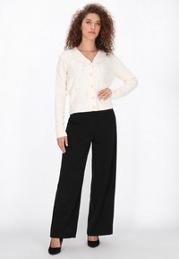 Valkoinen kaapelikudottu neuletakki, jossa on v-aukkoinen kaula-aukko ja nappikiinnitys, yhdistetty mustiin leveisiin lahkeisiin housuihin ja mustiin korkokenkiin, valkoista taustaa vasten.