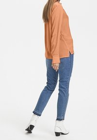 Blusa a maniche lunghe di un vivace arancione solido con polsini abbottonati, abbinata a jeans skinny blu e stivali alla caviglia bianchi con tacco, che mettono in risalto un design elegante.