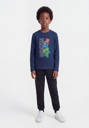 Pojke står klädd i en marinblå Ninjago långärmad tröja, svarta byxor och vita sneakers mot en enfärgad vit bakgrund.