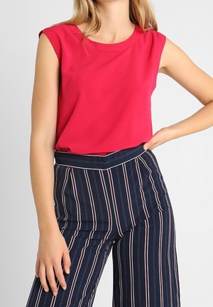 Femme portant un haut sans manches rouge vif rentré dans un pantalon bleu marine taille haute avec des rayures verticales blanches et rouges, main sur la hanche.