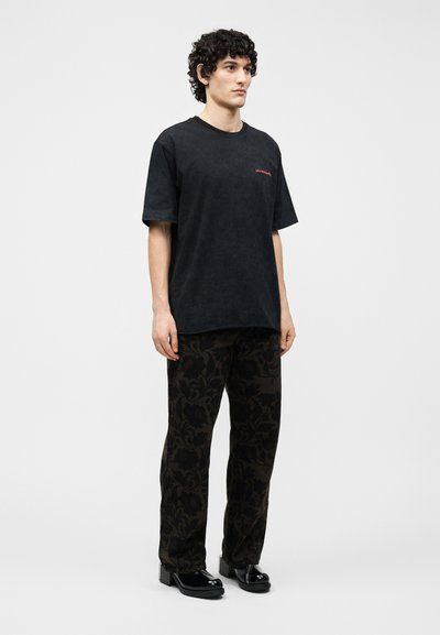 Άνδρας με κατσαρά μαύρα μαλλιά στέκεται φορώντας μαύρο υπερμεγέθες T-shirt, σκούρο παντελόνι με φλοράλ μοτίβο και γυαλιστερά μαύρα παπούτσια με πλατφόρμα, μπροστά σε λευκό φόντο.