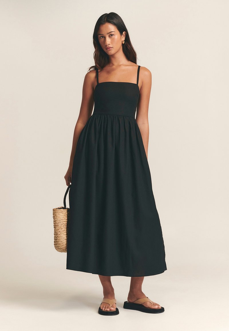 Vestido maxi negro sin mangas con cuerpo ajustado y falda fruncida. Lleva sandalias planas y sostiene un bolso de paja tejido.