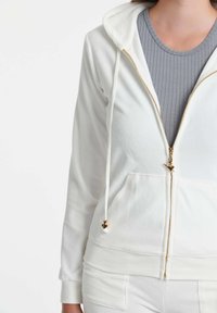 Bjela zip-up dukserica s prednjim džepom, zlatnim rajsferšlusom i vezicama, nosi se preko sive rebraste majice. Glatki materijal od pamuka i poliestera.
