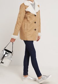 Vaate tan faux suede -takki valkoisella tekokarvatukalla ja -hihoilla, viisi nappia, yhdistettynä tummiin farkkuihin, hopeisiin kengännauhoihin ja metalliseen käsilaukkuun.