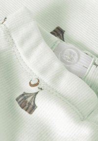 Close-up van lichtgroene geribbelde stof met bruine patronen, met een doorzichtige knoop genaaid op een witte verstelbare elastische band.