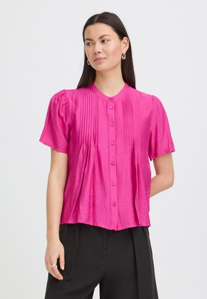 Femme portant un chemisier rose vif à manches courtes boutonné avec des plis, associé à un pantalon noir, debout devant un fond uni.