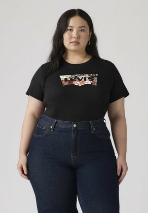 Frau mit langen dunklen Haaren, die goldene Creolen, ein schwarzes florales Levi's T-Shirt und dunkelblaue, hoch taillierte Jeans vor einfarbigem Hintergrund trägt.