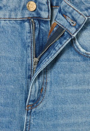 Mujer de pie que lleva una camisa vaquera azul oscuro con un pañuelo estampado en el cuello, vaqueros de pierna ancha azul claro y zapatos marrones sobre un fondo liso.