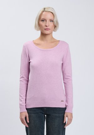 Femme blonde portant un pull violet clair à manches longues et un jean foncé, debout devant un fond uni gris clair.