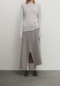 Pull en tricot gris clair avec un col rond, associé à une longue jupe portefeuille grise avec une fente diagonale à l'avant. Des mocassins noirs complètent la tenue.