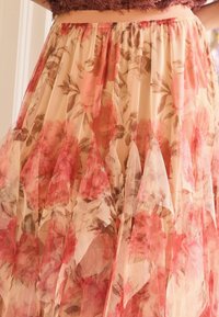 Floraler Maxirock aus transparentem, mehrlagigem Stoff in sanftem Beige mit rosa und roten Rosenmustern, mit Faltenwurf und strukturierten Details.