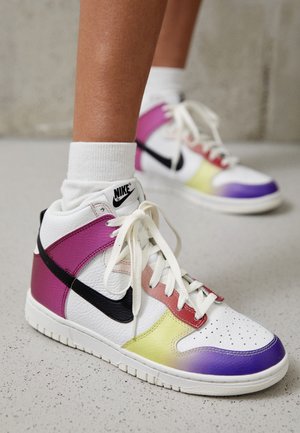 Białe wysokie sneakersy Nike z czarnym logo, fioletowymi, żółtymi i czerwonymi akcentami, noszone z białymi skarpetkami na szarej podłodze.