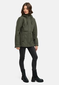 Femme portant une veste à capuche olive, un jean skinny noir et des bottines noires à semelles épaisses, se tenant les yeux fermés et la tête légèrement baissée.