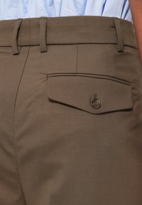 Hnedé nohavice majú plochý predok s pútkami na opasok a dekoratívnu vrecko s klopou a gombíkovým uzáverom. Hladká textúra, strihovaný dizajn.