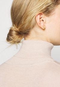 Blond kvinna med håret i en låg knut, iförd en beige polotröja och en liten guldring i höger öra.