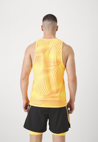 Żółty tank top bez rękawów z czerwonym wzorem fal, wykonany z lekkiego materiału. W połączeniu z czarnymi szortami. Prosty design, bez widocznych logo.