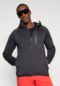 Melns jaku ar rāvējslēdzēju, ar pieguļošu dizainu, teksturētu audumu, regulējamu kapuci, apmali kabatu un rāvējslēdzēja aizdari, apvienota ar spilgti sarkanām biksēm.