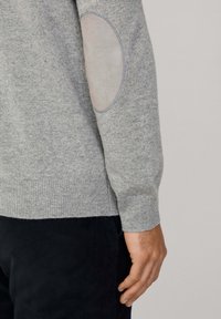 Szary sweter dzianinowy z prążkowanym dołem i ściągaczami, z jasnoszarymi zamszowymi łatkami na łokciach. Zbliżenie na rękaw i dłoń.