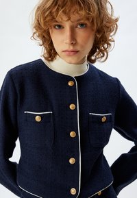 Giacca blu strutturata con bottoni dorati, rifiniture bianche e due tasche frontali. Presenta un design aderente e un collo rotondo.