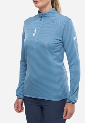 Femme portant un haut de sport à manches longues texturé bleu clair avec une fermeture éclair à l'avant et des logos "Millet" blancs sur la poitrine et la manche.