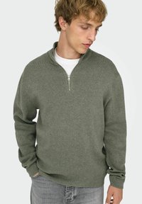 Olijfgroene trui met ritssluiting, een gestructureerd breipatroon, geribbelde manchetten en een opstaande kraag, gedragen over een wit T-shirt.