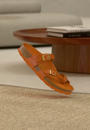 Narancssárga szandál két állítható pánttal, arany csatokkal, texturált parafa talpbéléssel és gumitalppal. Egy texturált felület felett lebeg.