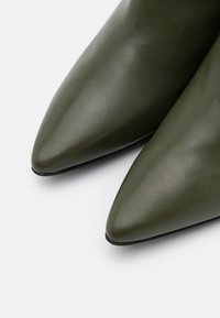 Spitz zulaufende Schuhe aus dunkelolivgrünem Leder. Glatte Textur mit elegantem Design und schwarzen Sohlen. Sichtbare Nahtdetails entlang der Kanten.