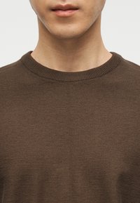 Κοντινό πλάνο ενός ατόμου που φοράει καφέ πλεκτό πουλόβερ με στρογγυλή λαιμόκοψη, εμφανίζοντας τον λαιμό, το κάτω μέρος του προσώπου και το πάνω μέρος του στήθους.