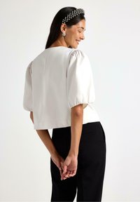 Hvid cropped top med puffærmer, glat tekstur, rund halsudskæring, parret med sorte skræddersyede bukser. Håret prydet med et perle-hovedbånd.