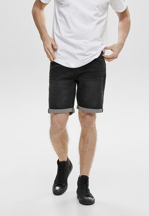 Schwarze Denim-Shorts mit aufgerollten Säumen, einer geraden Passform und dezenten Stickdetails, kombiniert mit einem schlichten weißen T-Shirt und schwarzen Sneakers.