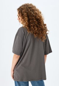Camiseta gris de gran tamaño con mangas cortas, hecha de una tela suave. Presenta un corte relajado y costuras simples. Se lleva con unos pantalones vaqueros azules.