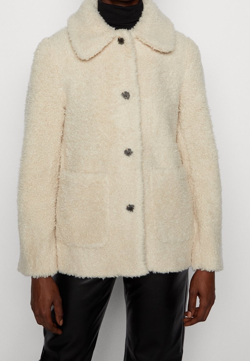Veste boutonnée en sherpa couleur crème avec grand col, poches plaquées frontales, portée sur un col roulé noir et un pantalon noir.