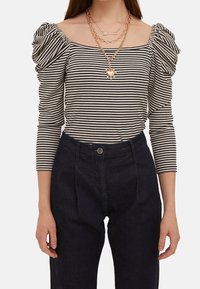 Pruhovaná blůza s odhalenými rameny a nafouknutými rukávy v černé a bílé barvě, kombinovaná s tmavými kalhotami. Nahrdelníky ze zlata v několika vrstvách zvýrazňují výstřih.