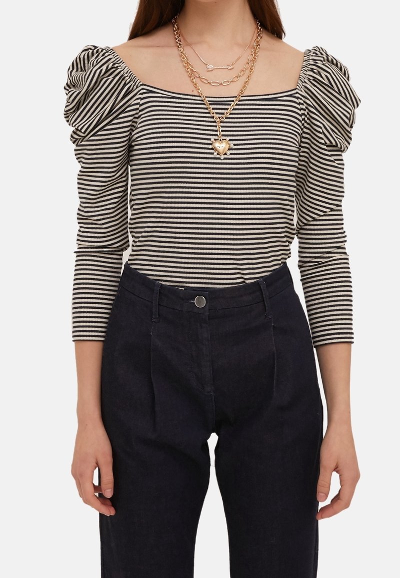 Pruhovaná blůza s odhalenými rameny a nafouknutými rukávy v černé a bílé barvě, kombinovaná s tmavými kalhotami. Nahrdelníky ze zlata v několika vrstvách zvýrazňují výstřih.