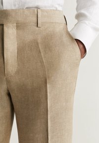 Beżowe lniane spodnie z płaskim przodem, bocznymi kieszeniami i dopasowanym krojem, w połączeniu z białą koszulą z długim rękawem.