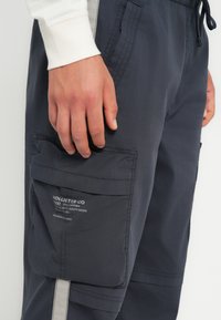 Tumši pelēki kravas bikses, izgatavotas no viegla auduma. Tām ir liels sānu kabata ar zīmolu, kontrastējošs šuvums un auklas josta.