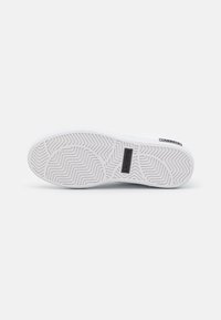 Balts gumijas sneaker zole ar teksturētu rakstu. Satur melnu zīmola etiķeti ar tekstu "LACOSTE," nodrošinot saķeri uz dažādām virsmām.