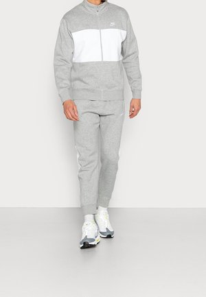 Homme portant une veste zippée Nike grise et blanche, un pantalon de survêtement gris assorti avec des bandes blanches sur les côtés, et des baskets blanches, debout sur un fond uni.