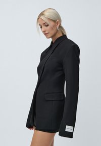 O femeie blondă îmbrăcată într-un sacou negru ajustat, cu mâneci lungi și o etichetă albă pe manșetă, pozând pe un fond deschis simplu.